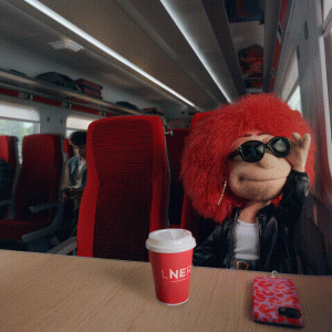 LNER's red-headed muppet style mascot sits on a train, with her phone and a coffee on the table in front of her, looking out the window wearing sunglasses and smiling.