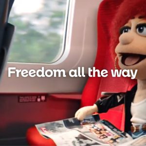 LNER's red-headed puppet mascot sits on a train., smiling and reading a magazine.